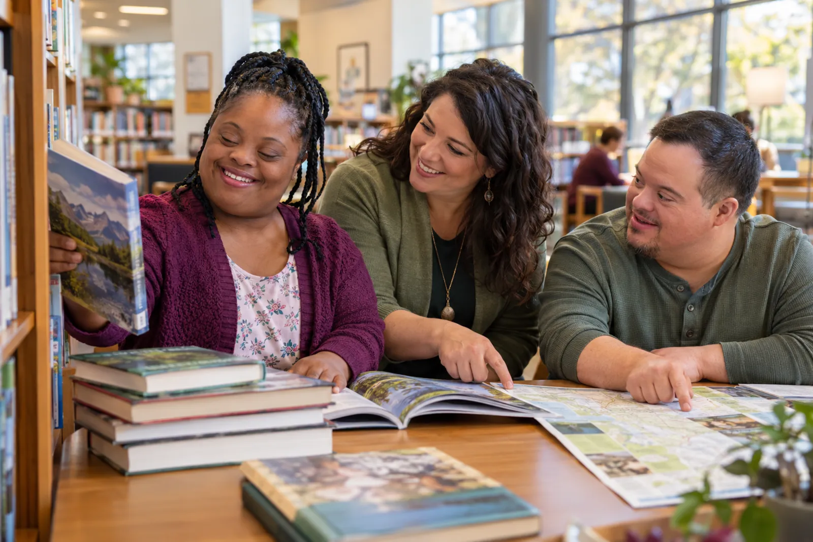 Two adults choose books and point out places on a map at a library table while a support professional joins in planning an outing.